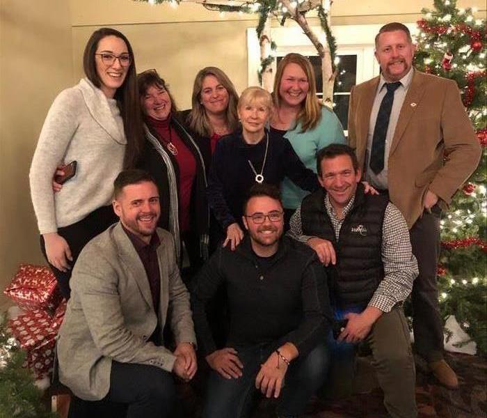 Group of people in business casual clothes at a holiday party with a Christmas tree to the right, presents to the left.