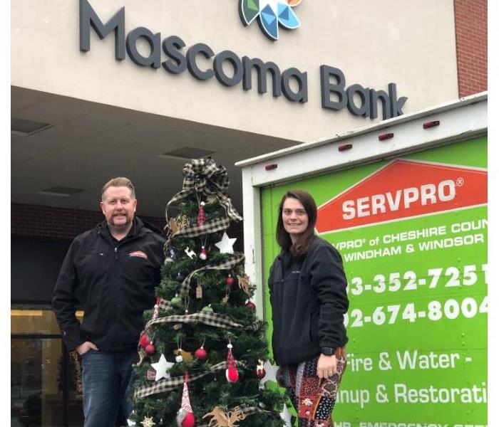 Two SERVPRO employees standing on a SERVRPO truck gate with a decorated Christmas tree in front of a bank.