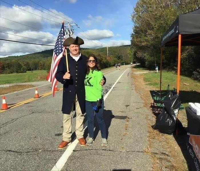 man in Patriot costume with American flag over shoulder stands nest to woman in green shirt and sunglasses outside on road.